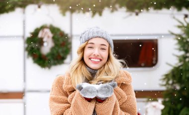 Tüylü palto ve örgü şapkalı genç bir kadın taze yağmış kar tutarken parlak bir şekilde gülümsüyor. Kış sahnesinde şenlikli dekorasyonlar ve hafif kar yağışı yer alıyor, neşeli bir atmosfer yaratıyor..