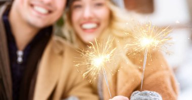 Neşeli bir çift, ellerinde yüzlerini aydınlatan kıvılcımlar tutarak dışarıda poz veriyor. Şenlikli bir kış akşamının tadını çıkarıyorlar. Kahkahalarını ve sıcacık kıyafetlerini paylaşıyorlar..