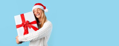 Muhteşem bir bayan büyük hediye kutusunu tutuyor ve sarılıyor. Mavi stüdyo arka planında Noel Baba şapkası takıyor. Noel hediyeli mutlu kadın Noel 'i ya da yeni yılı kutlamaya hazır, boşluğu kopyala