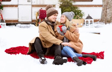 A young couple sits on a red blanket in the snow, sharing a warm moment together. Surrounded by holiday decorations, they smile at each other as snowflakes gently fall around them. clipart