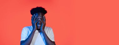 Genç bir siyah adam parlak turuncu bir arka fonun önünde duruyor, yüzünde elleri ile şaşırmış bir ifade sergiliyor. Günlük kıyafetleri, etrafındaki canlı renklerle çelişiyor..