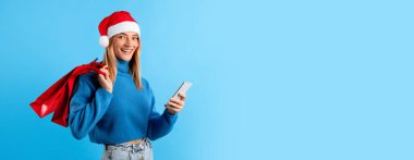 Noel Baba şapkalı genç Avrupalı kadın elinde alışveriş çantaları ve akıllı telefon kullanıyor. İnternetten alışveriş yapıyor. Xmas satışları ise mavi stüdyo arka planında duruyor.