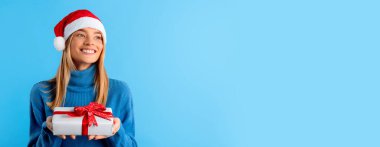 Noel hediyesi. Mutlu kadın hediye kutusunu tutuyor ve boş alana bakıyor mavi stüdyo arka planında poz veriyor, panorama. Kış tatili kutlaması, yeni yıl reklamı