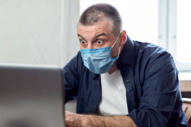 Coronavirus ve Kriz. Şok olmuş adam bilgisayar başında koruyucu maske takarak çalışıyor. Covid-19 salgını sırasında evde oturuyor..