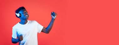 Kulaklık takan Afrikalı Amerikalı genç adam müzik dinlerken neşeyle dans ediyor, parlak kırmızı bir arka plana karşı kaygısız bir tavır sergiliyor..