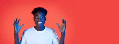 Genç bir siyah adam geniş bir alanda gülümsüyor ve heyecan içinde ellerini kaldırıyor, canlı kırmızı bir arkaplanda. İfade bir neşe ve coşku iletir..