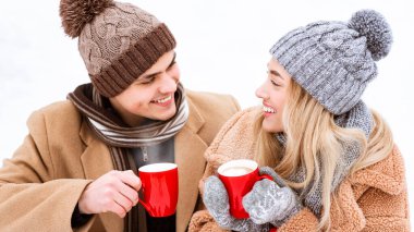 Sıcak kışlık giysiler giymiş genç bir çift, karlı bir manzaranın ortasında otururken sıcak içecekleri mutlulukla paylaşıyorlar. Neşeli ifadeleri soğuk hava cazibesini yansıtıyor..