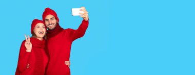 Kışlık şapkalı ve eşarplı mutlu çift akıllı telefondan selfie çekiyor, neşeli genç adam ve kadın cep telefonuyla fotoğraf çekiyor, mavi stüdyo arka planında tek başına duruyor, uzay kopyaları yapıyor.