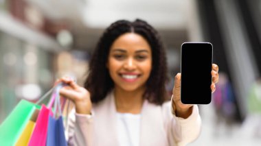 İndirimli uygulama. Gülümseyen siyah kadın alışveriş merkezinde alışveriş yaptıktan sonra akıllı telefonu siyah ekranla gösteriyor, seçici odak ve boş alan ile görüntüyü taklit ediyor