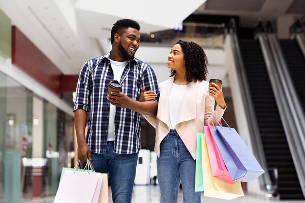 Любители шоппинга. Веселая черная пара прогулки с вынос кофе и сумки в торговом центре, наслаждаясь сезонные продажи и скидки, счастливые африканские супруги делать покупки вместе, бесплатное пространство