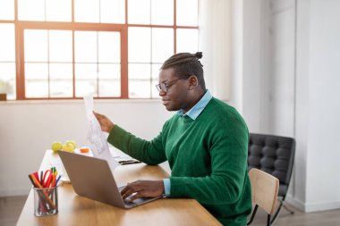 Gözlüklü siyah bir adam parlak bir ofis yerinde ahşap bir masada oturur. Ofis malzemeleri ve fabrikalarla çevrili bir dizüstü bilgisayar üzerinde çalışırken belgeleri incelemeye odaklanmış durumda..