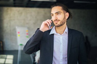 Çağdaş bir ofiste dururken, koyu takım elbiseli genç bir adam telefonuyla konuşur. Çevrede parlak bir ışıklandırma ve arka planda bir beyaz tahta var.