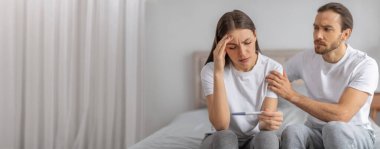 Bir kadın hamilelik testi yaparken endişeli görünürken, eşi rahatlık sunuyor. Yumuşak ışıklı bir odada birlikte bir yatakta oturuyorlar, endişe dolu bir anı paylaşıyorlar..