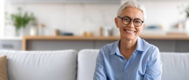 Gri saçlı ve gözlüklü neşeli yaşlı bir kadın, oturma odasındaki yumuşak bir kanepede rahatça otururken neşe saçıyor. Bitkiler ve ışıkla dolu..