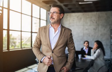 İyi giyimli bir işadamı, çağdaş bir ofiste pencerenin kenarında duruyor ve bir tartışmayla meşgul oluyor. Bir masada oturan iki meslektaşı, toplantı sırasında odaklanmış ve dikkatli görünüyorlar..