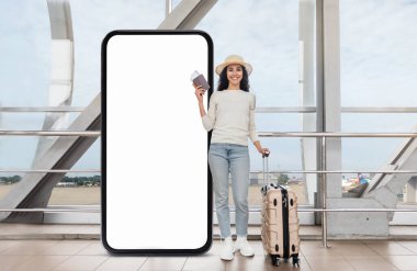 Havaalanı terminalinde elinde akıllı telefon ve seyahat belgeleriyle gülümseyen bir gezgin, boş ekranlı dev bir telefonun yanında duruyor. Yanında bir bavul var, yolculuk için hazır..