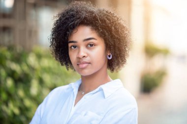 Genç bir kadın rahat bir tavırla beyaz gömlek giyerken kıvırcık saçlarını gösteriyor. Arkaplanda sıcak ve davetkar bir atmosfer yaratan yeşillikler var..