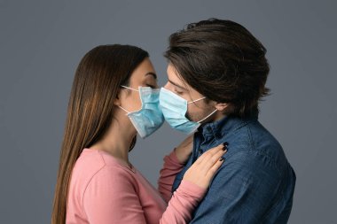 Romantik Karantina. Birbirlerine aşık genç bir çift koruyucu tıbbi maskelerle öpüşüyorlar, açık stüdyo arka planında duruyorlar, boş yer var.