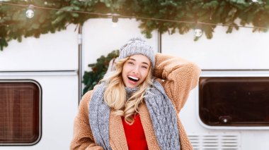 Rahat bir palto giyen ve örgü ören genç bir kadın açık havada gülümsüyor. Karlı bir arka planda, şenlikli süslemelerle süslenmiş eski bir römorkun yanında duruyor..