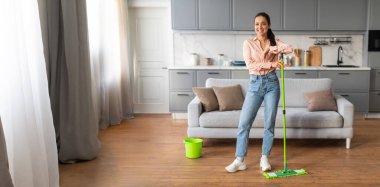 Genç bir kadın elinde paspasla güvenle ayakta duruyor, modern mutfağının ahşap zeminini ve yaşam alanını temizlerken gülümsüyor. Yakınlarda yeşil bir kova var, temizlik görevini gösteriyor..