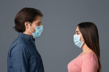 Sosyal Mesafe Kavramı. Tıbbi maskeli genç adam ve kadının yan görüntüsü. Işık stüdyosunun arka planında, boş alanda yüz yüze duruyorlar.