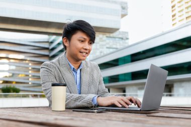 A young Asian man, dressed in a stylish blazer, is focused on his laptop while seated at a wooden table outside a contemporary building under the afternoon sun. A cup of coffee sits beside him. clipart