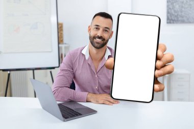 Dostça gülümseyen bir adam parlak bir ofiste boş bir akıllı telefonu tutuyor. Onun yanına bir dizüstü bilgisayar yerleştiriliyor, dijital aktiviteler için uygun bir çalışma alanı ortamı yaratıyor.