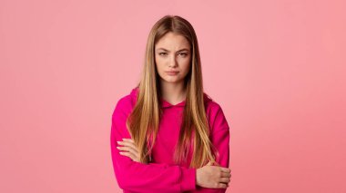 Güvensizlik konsepti. Genç kız kollarını kavuşturup kameraya bakıyor. Pembe arka planda izole edilmiş, stüdyo çekimi, boş alan.