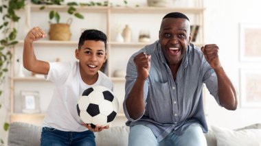 Siyah çocuk büyükbabasıyla televizyonda futbol maçı izliyor ve elinde futbol topuyla tezahürat yapıyor, evde birlikte vakit geçiriyor.