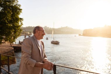 Güneş batarken, gülümseyen bir adam nehir kenarındaki korkuluklara yaslanır ve suyun ve çevrenin üzerine sıcak bir parıltı saçar. Tekneler yavaşça uzaklardan geçiyor..