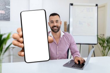 Gülümseyen bir adam modern bir ofiste masada oturuyor, elinde boş bir akıllı telefon ekranı var. Dizüstü bilgisayarla çalışırken, dost canlısı ve teknolojiden anlayan bir atmosferi ön plana çıkarıyor..
