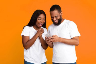 Heyecanlı siyah çift cep telefonu ekranına bakıyor, yeni bir uygulama kullanıyor, sarı turuncu stüdyo arka planı var. Neşeli Afro-Amerikan erkek ve kadın akıllı telefon kullanıyorlar, eğlence konsepti.
