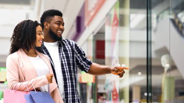 Tüketim kavramı. Mağazada birlikte alışveriş yapan mutlu siyah eşler, vitrindeki yeni kıyafetleri gösteren gülümseyen genç adam, alışveriş merkezinde alışveriş yapan Afrikalı Amerikalı çift,