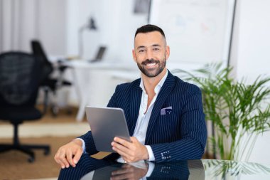 Şık çizgili takım elbise giymiş kendine güvenen bir adam çağdaş bir ofiste masa başında oturuyor. Elinde tablet tutarken gülümsüyor. Etrafı temiz ve düzenli bir çalışma alanıyla çevrili..