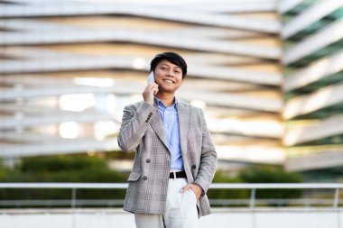 Genç bir Asyalı, gün ışığında çağdaş bir mimari yapının dışında telefonla konuşurken kendine güvenen bir takım elbiseyle ayakta duruyor..