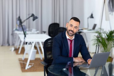 Takım elbiseli bir adam modern bir ofiste cam bir masada oturuyor. Minimalist mobilyalar ve kapalı alan bitkileriyle çevrili bir dizüstü bilgisayar kullanarak gülerek verimli bir atmosfer yaratıyor..