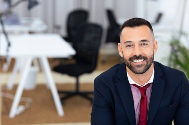 Düzgün sakallı ve kısa saçlı iyi giyimli bir adam çağdaş bir ofiste otururken sıcak bir şekilde gülümsüyor. Arkasında şık ofis mobilyaları ve parlak bir atmosfer var..