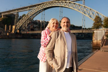 Nehir kıyısında bir çift yan yana durur, neşeyle gülümserler. Çarpıcı köprü arka planda açık mavi gökyüzüne karşı kemerler oluşturarak nişanları için pitoresk bir ortam oluşturuyor..