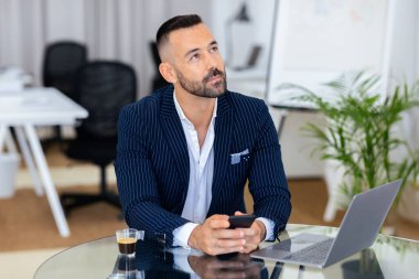 Şık, ince çizgili bir takım elbise giyen adam, çağdaş bir ofiste cam bir masada oturuyor, yanında bir laptop ve kahve varken telefonuna odaklanıyor..