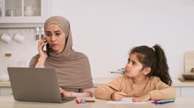 Başörtüsü takan bir anne, mutfak masasında otururken telefon konuşmasına odaklanır. Onun yanında, kızı renkli kalemlerle resim çiziyor, merak ve endişe gösteriyor..