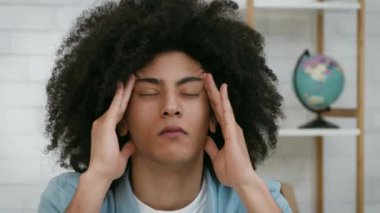Kıvırcık saçlı siyahi adam içeride oturuyor, kafasını tutarken gözle görülür bir şekilde stresle boğuşuyor. Arkaplanda bir küre ve modern bir dekor yer alıyor..