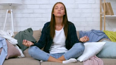 Genç bir kadın kanepede bağdaş kurmuş oturuyor. Gözleri meditasyonda kapalı. Etrafı dağınık kıyafetlerle çevrili..