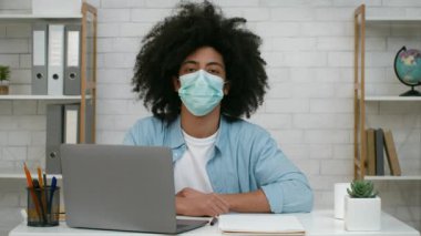 Genç bir adam dizüstü bilgisayarla masada oturuyor, maske takıyor ve kameraya bakıyor. İyi organize edilmiş bir çalışma alanından online bir derse odaklanmış ve meşgul görünüyor..