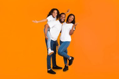Aileyle vakit geçirmek eğlencelidir. Heyecanlı Afrikalı erkek, kadın ve kız tüm vücut uzunluğu sarı turuncu stüdyo duvarında izole bir şekilde gülüyor ve poz veriyor. Neşeli baba kızını sırtında taşıyor