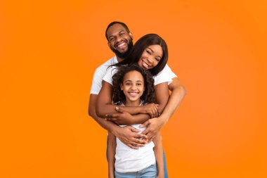 Sağlıklı Aile İlişkisi. Arkasından karısına ve kızına sarılan neşeli Afro-Amerikalı adamın portresi sarı turuncu stüdyo duvarında dikilip kameraya bakıyor.