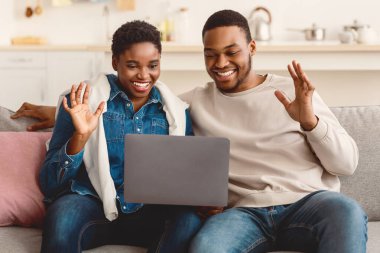 Çevrimiçi İletişim. Gülümseyen Afro-Amerikan erkek ve kadın arkadaşlarıyla dijital görüntülü sohbet yapıyorlar. Laptop kullanıyorlar, web kameralarına el sallıyorlar, oturma odasındaki kanepede oturuyorlar.