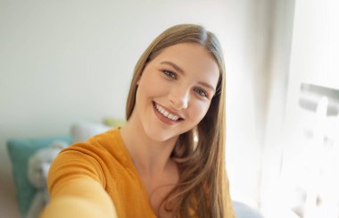 Uzun saçlı genç bir kız neşeli bir selfie çekmek için kamerasını tutuyor. Güneş ışığı pencereden akar ve odanın sıcak atmosferine sıcaklık katar..