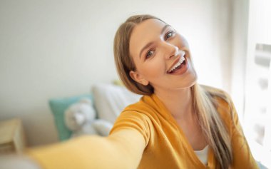 Uzun saçlı neşeli bir genç kız parlak, davetkar bir odada selfie çekiyor. Güneş ışığı pencereden süzülerek sıcak bir atmosfer yaratır..