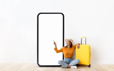 Hardal renkli bluzlu neşeli bir kadın ve büyük boş bir telefon ekranını göstererek gülümsüyor. Yerde yanında parlak sarı bir bavul duruyor, seyahat temalı bir atmosfer yaratıyor..