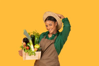 Wearing a green shirt and apron, a smiling black woman with a straw hat showcases a wooden crate filled with vibrant vegetables. The bright yellow background enhances her joyful expression. clipart